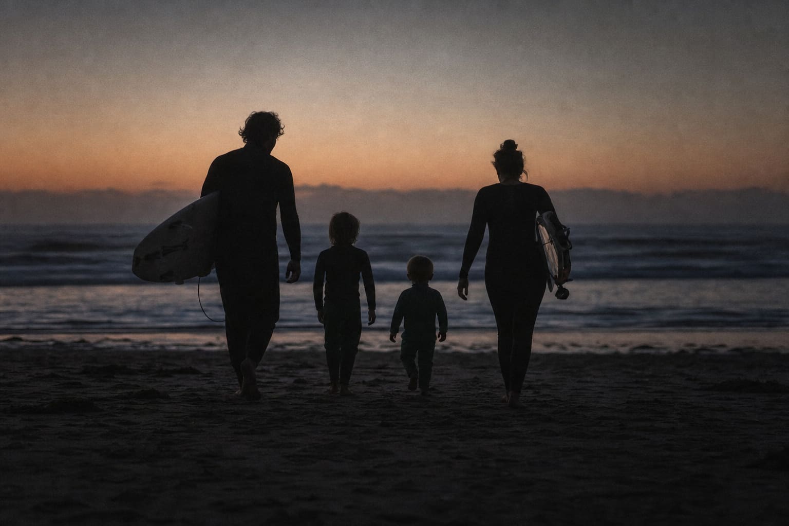 A family walking toward the ocean at dusk carrying surfboards