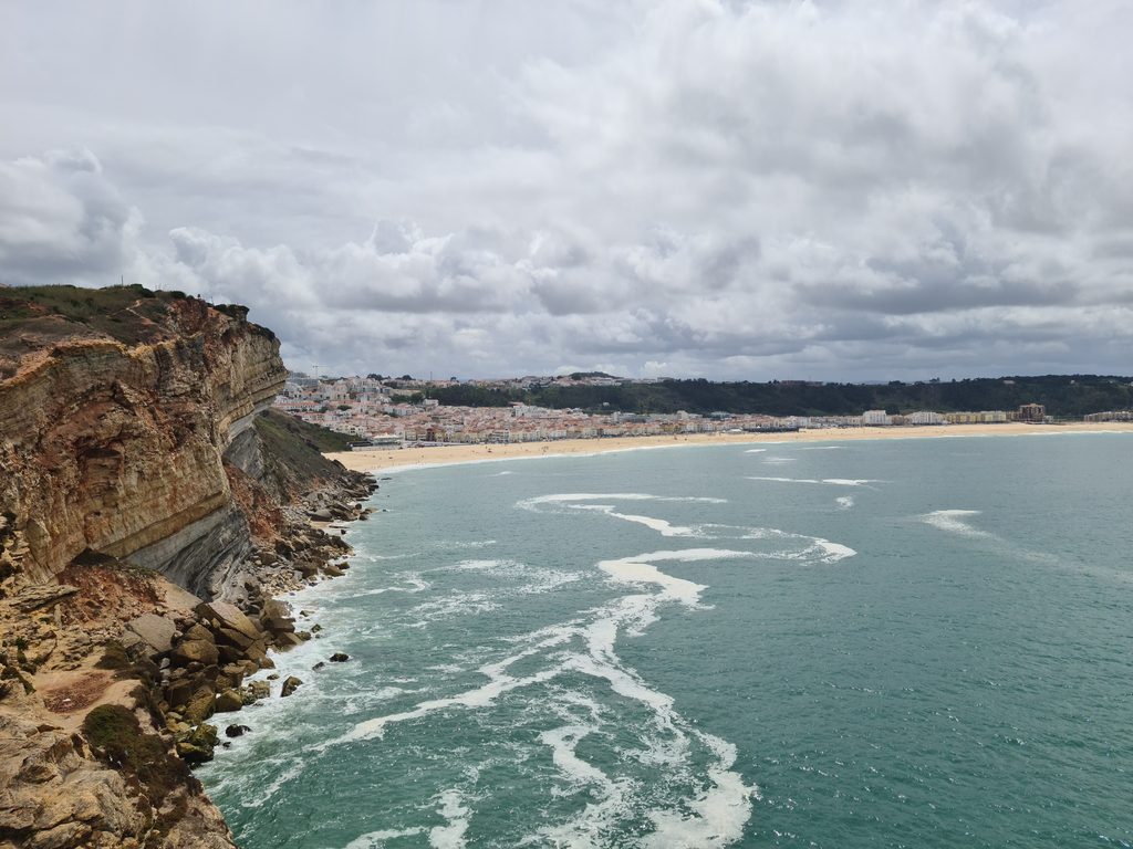 A wide sandy bay in front of a coastal town, with cliffs and a clifftop neighbourhood in the background
