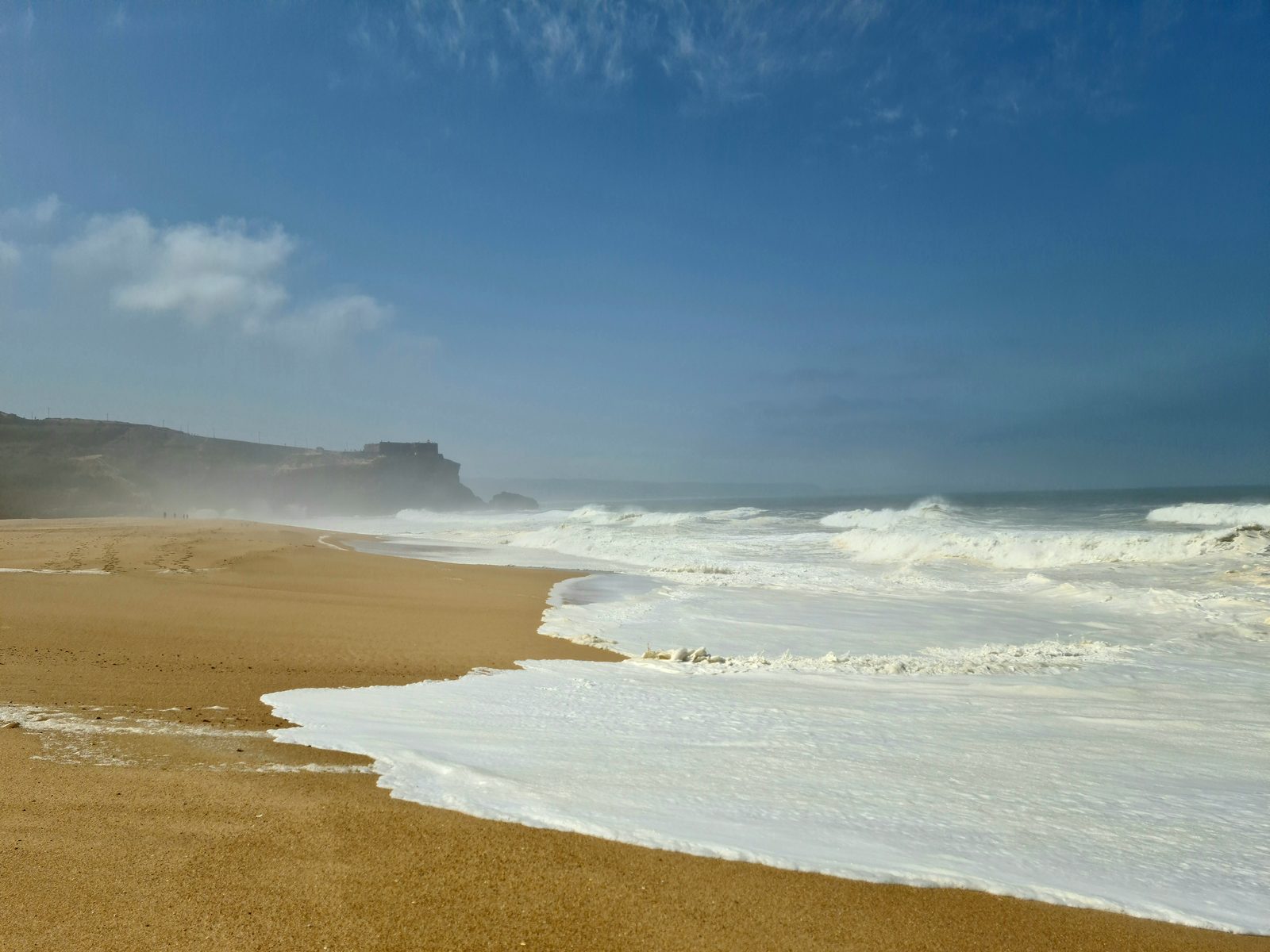 Large white-water waves breaking along a sandy beach beneath cliffs near Nazaré
