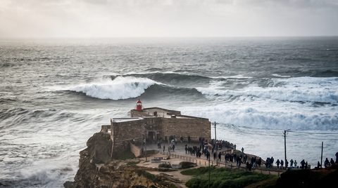 The Forte de São Miguel Arcanjo lighthouse on its cliff, with a big wave breaking beyond and spectators gathered on the headland