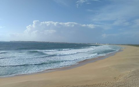 A wide, empty stretch of sand at Praia da Consolação with rolling whitewater and the Peniche headland in the distance